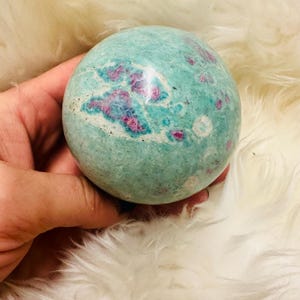 Könnte beinhalten: Eine runde, polierte Edelstein-Kugel. Der Stein hat eine gesprenkelte türkisfarbene Farbe mit Flecken in leuchtendem Pink und Weiß. Die Kugel wird in einer Hand vor einem flauschigen, weißen Hintergrund gehalten.