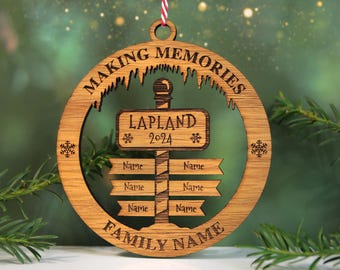 May include: Wooden Christmas ornament with the words "Making Memories" and "Family Name" engraved. The ornament features a signpost design with "Lapland 2024" and spaces for names. It hangs from a red and white string.