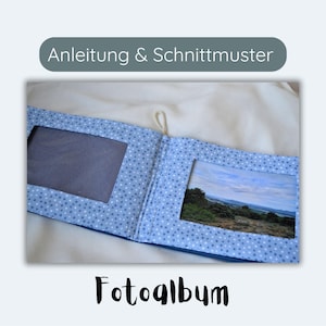 May include: Open photo album with blue fabric cover and white polka dots. The album has two photo windows, one empty and one with a landscape photo. The text "Fotoalbum" is written in black script.