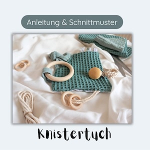 Puede incluir: Un juguete para bebé de tela de tejido de gofre color turquesa con un anillo de madera, una bola de madera y una cuerda blanca. El juguete está sobre una superficie blanca con el texto "Anleitung & Schnittmuster" y "Knistertuch" visible.