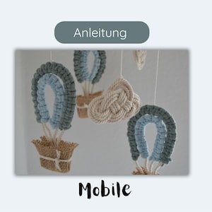Könnte beinhalten: Handgefertigtes Mobile mit Makramee-Heißluftballons in Blau- und Cremetönen. Die Ballons haben eine gewebte Korbbasis. Das Wort "Mobile" steht unten in einer Schriftart. Das Wort "Anleitung" steht oben.