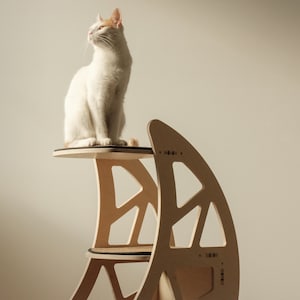 May include: A white cat with orange markings sits on a modern, light-coloured wooden cat tree. The cat tree has multiple levels and a unique geometric design, providing a stylish space for feline relaxation and play.