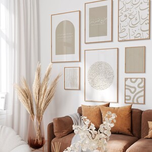 Op de afbeelding: Een woonkamerinterieur met een bruine leren bank, een witte fauteuil en een salontafel. De muren zijn versierd met een galerijmuur van ingelijste prints met abstracte ontwerpen en Arabische kalligrafie. Een vaas met gedroogd pampasgras en een vaas met witte bloemen staan op de salontafel.
