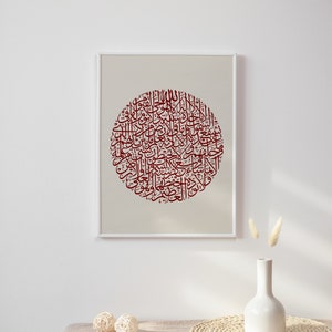 May include: A framed print of a circular design featuring intricate Arabic calligraphy in a deep red color on a beige background. The print is displayed on a white wall with a vase of dried flowers and a woven placemat on a beige table.