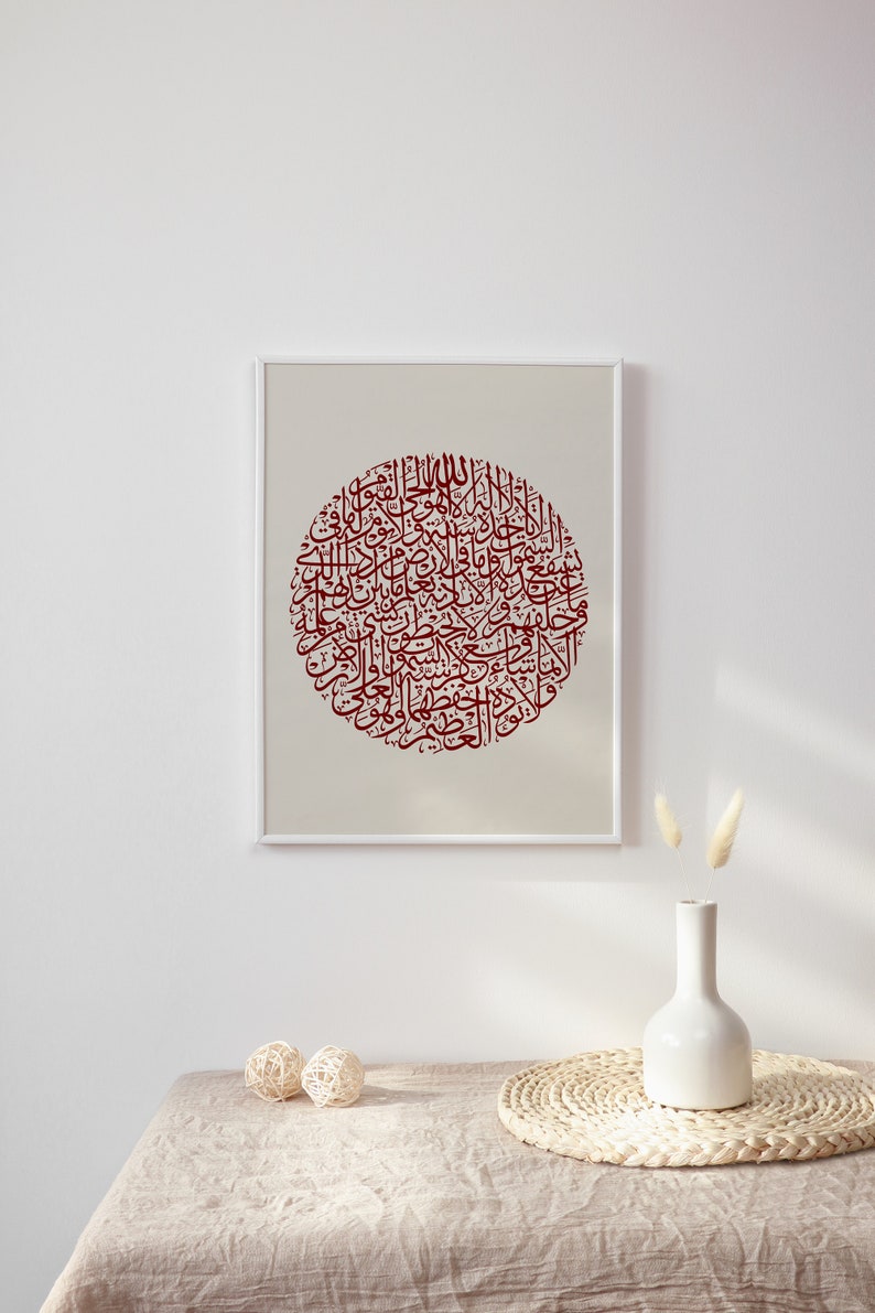 May include: A framed print of a circular design featuring intricate Arabic calligraphy in a deep red color on a beige background. The print is displayed on a white wall with a vase of dried flowers and a woven placemat on a beige table.