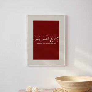 May include: A framed print with a red background and white Arabic calligraphy. The text translates to "Indeed with every difficulty comes ease".