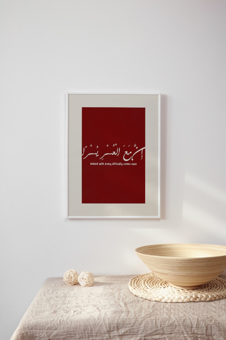 May include: A framed print with a red background and white Arabic calligraphy. The text translates to "Indeed with every difficulty comes ease".