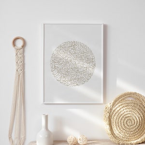 Op de afbeelding: Een ingelijst afdruk met een cirkelvormig ontwerp van Arabische kalligrafie in een lichtbruine kleur. De afdruk hangt aan een witte muur boven een tafel met een witte vaas, twee beige geweven ballen en een beige geweven tas.