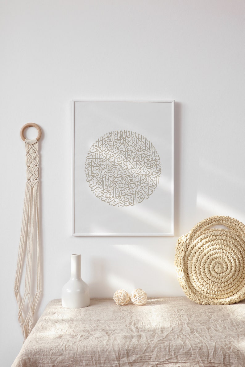 Op de afbeelding: Een ingelijst afdruk met een cirkelvormig ontwerp van Arabische kalligrafie in een lichtbruine kleur. De afdruk hangt aan een witte muur boven een tafel met een witte vaas, twee beige geweven ballen en een beige geweven tas.