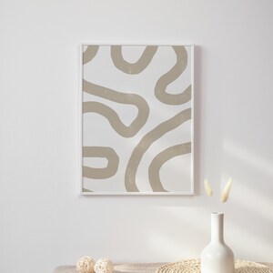Op de afbeelding: Een ingelijst abstract print met beige lijnen op een witte achtergrond, een witte vaas met gedroogd pampasgras en twee geweven ballen op een beige linnen tafelkleed.
