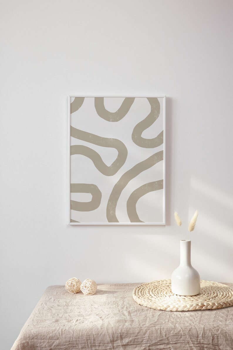 Op de afbeelding: Een ingelijst abstract print met beige lijnen op een witte achtergrond, een witte vaas met gedroogd pampasgras en twee geweven ballen op een beige linnen tafelkleed.