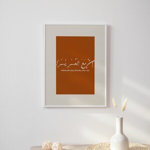 Peut inclure: Impression d'art encadr&eacute;e avec un fond orange br&ucirc;l&eacute; et une calligraphie blanche. Le texte se lit comme suit : &laquo; En effet, avec chaque difficult&eacute; vient la facilit&eacute; &raquo;. L'impression est affich&eacute;e sur un mur blanc au-dessus d'une table.