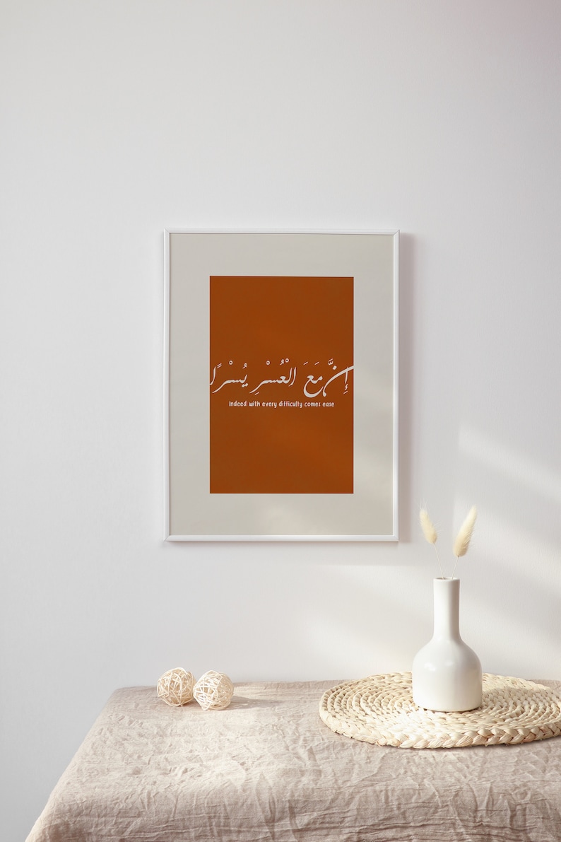 Peut inclure: Impression d'art encadr&eacute;e avec un fond orange br&ucirc;l&eacute; et une calligraphie blanche. Le texte se lit comme suit : &laquo; En effet, avec chaque difficult&eacute; vient la facilit&eacute; &raquo;. L'impression est affich&eacute;e sur un mur blanc au-dessus d'une table.