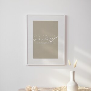 Op de afbeelding: Een ingelijst print met een beige achtergrond en witte Arabische kalligrafie die "Inderdaad, met elke moeilijkheid komt gemak" luidt. De print hangt aan een witte muur met een witte vaas en gedroogde bloemen ervoor.