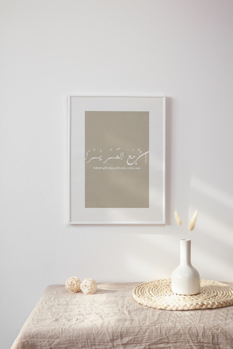 Op de afbeelding: Een ingelijst print met een beige achtergrond en witte Arabische kalligrafie die "Inderdaad, met elke moeilijkheid komt gemak" luidt. De print hangt aan een witte muur met een witte vaas en gedroogde bloemen ervoor.