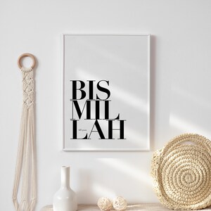 May include: A white framed print with the Arabic phrase "Bismillah" in black text. The print is hanging on a white wall above a beige table with a white vase, two beige balls, and a woven straw bag.