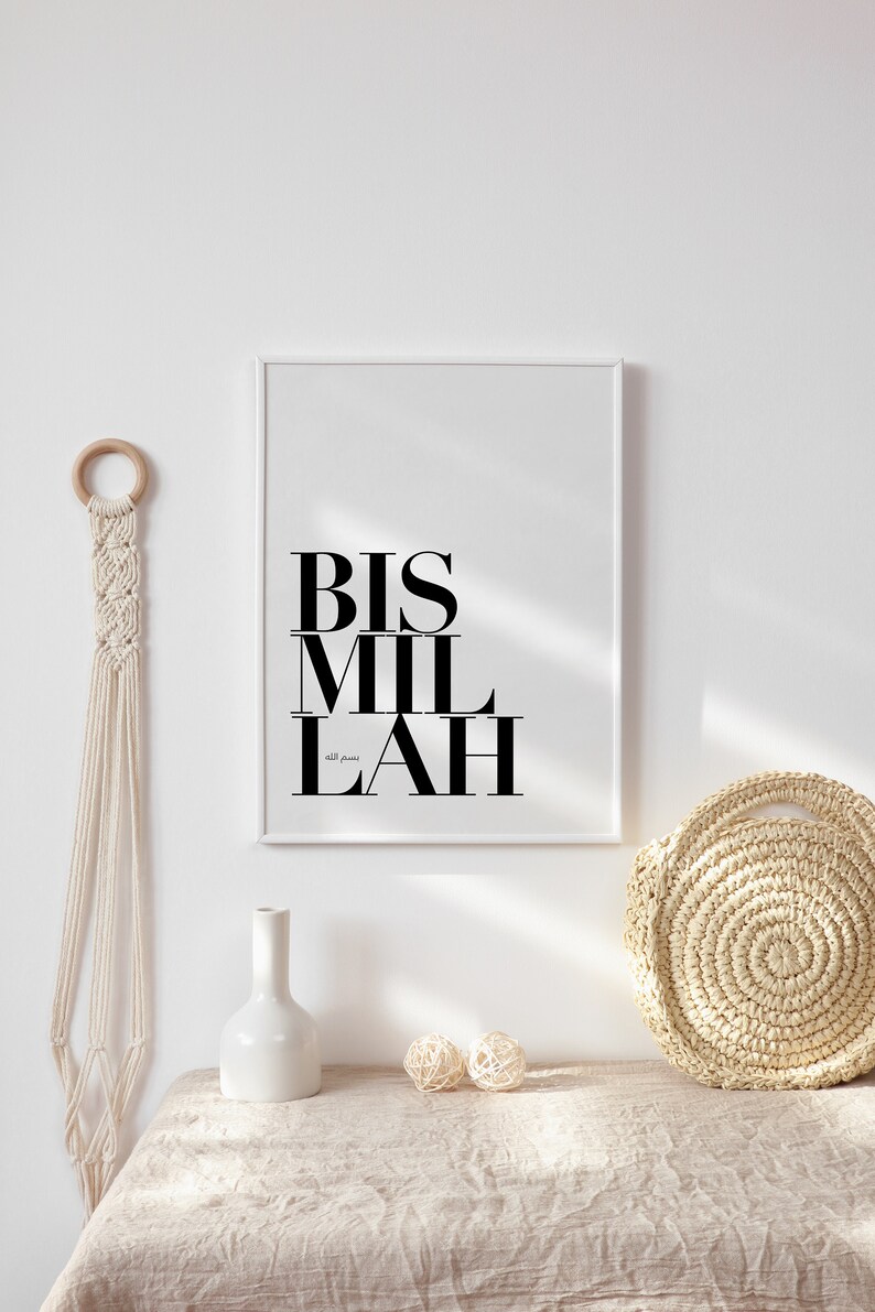 May include: A white framed print with the Arabic phrase "Bismillah" in black text. The print is hanging on a white wall above a beige table with a white vase, two beige balls, and a woven straw bag.