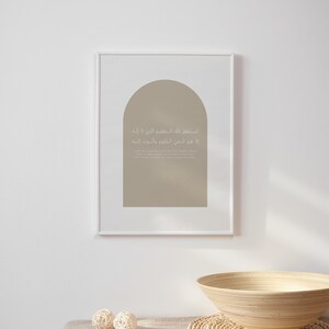 Op de afbeelding: Een ingelijst afdruk met een beige achtergrond en een boogvorm. De afdruk bevat Arabische tekst in wit, die vertaalt naar "In de naam van Allah, de Meest Barmhartige, de Meest Genadevolle." De afdruk hangt aan een witte muur boven een houten kom en een geweven mat.