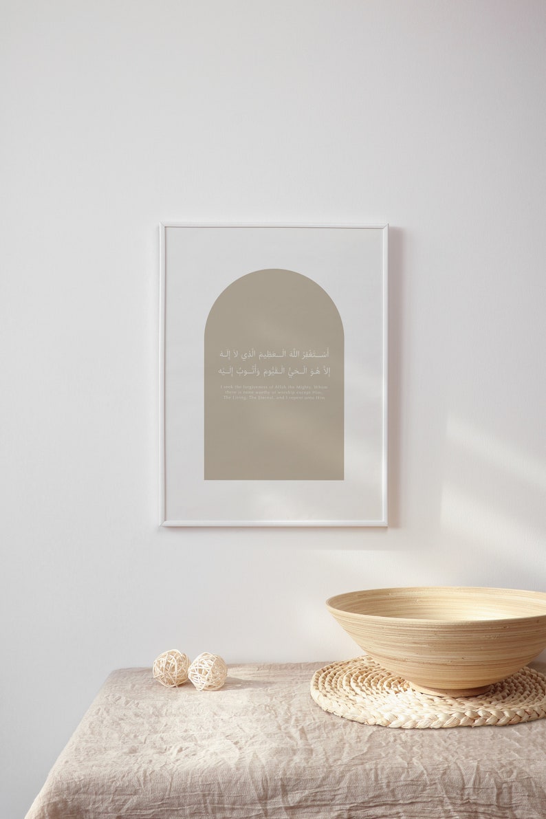 Op de afbeelding: Een ingelijst afdruk met een beige achtergrond en een boogvorm. De afdruk bevat Arabische tekst in wit, die vertaalt naar "In de naam van Allah, de Meest Barmhartige, de Meest Genadevolle." De afdruk hangt aan een witte muur boven een houten kom en een geweven mat.