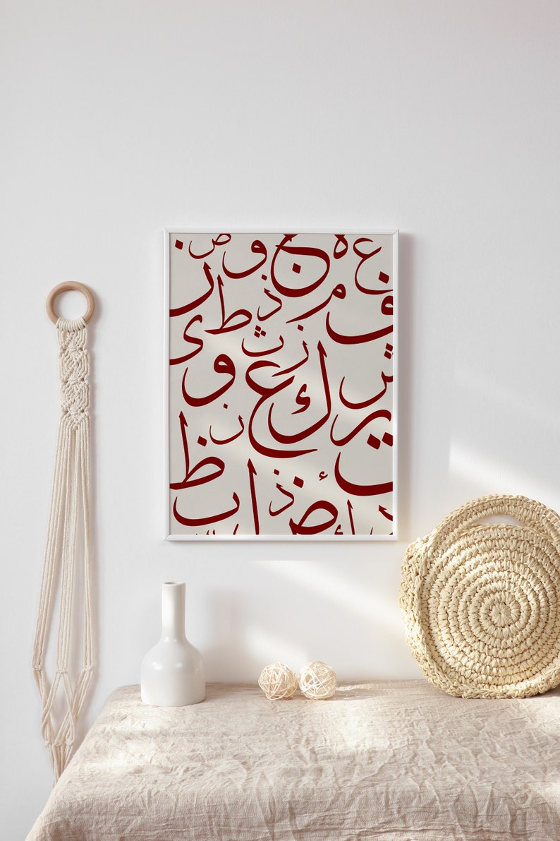 May include: A framed print with a white background and red Arabic calligraphy. The print is hanging on a white wall above a table with a white vase and a woven basket.