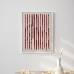 May include: A framed abstract print with vertical red lines on a white background. The print is hanging on a white wall above a table with a vase and decorative balls.