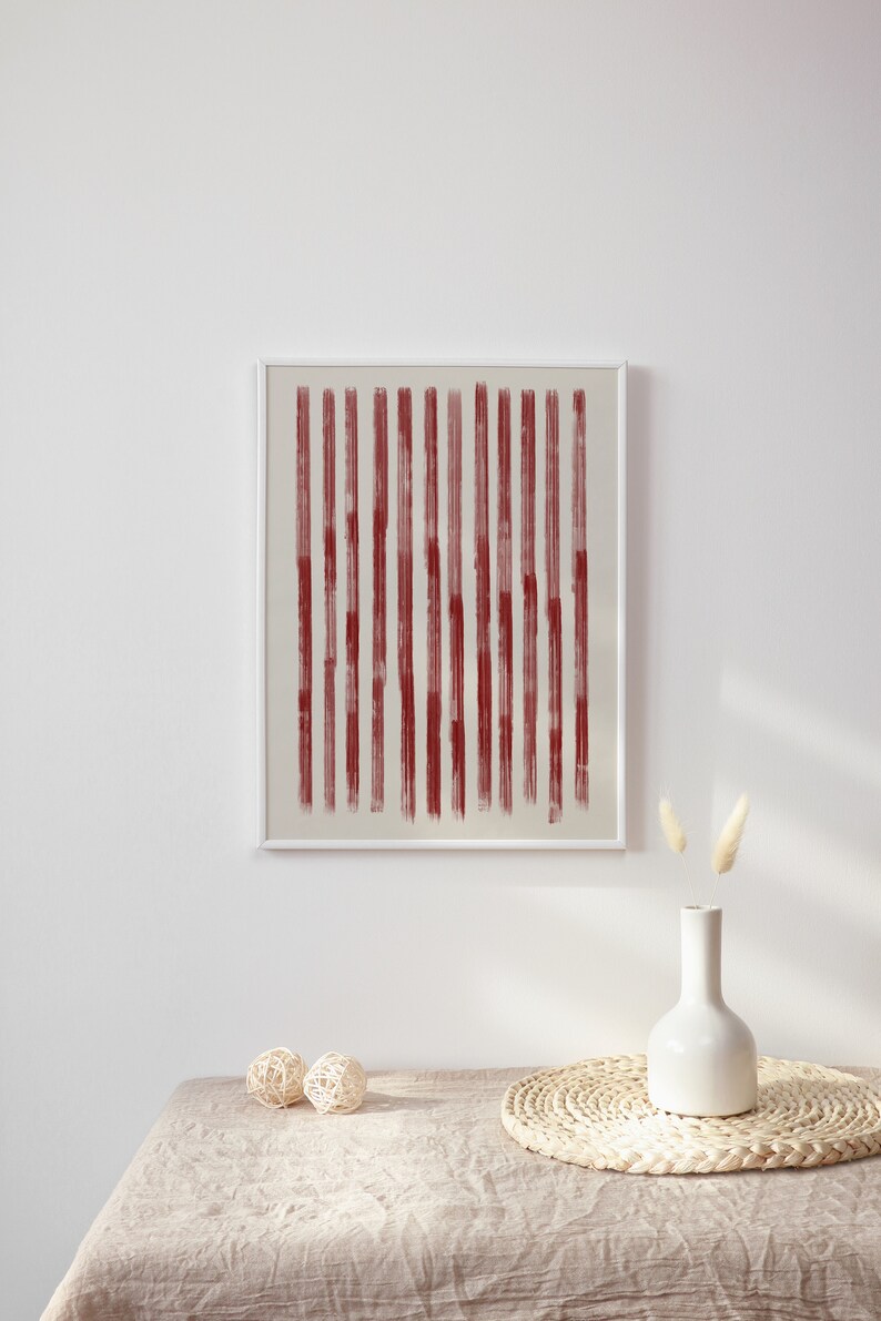 May include: A framed abstract print with vertical red lines on a white background. The print is hanging on a white wall above a table with a vase and decorative balls.