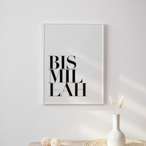 May include: A black and white print with the Arabic phrase "Bismillah" in a modern font style. The print is framed in a white frame and is hanging on a white wall. There is a white vase with dried grass on a table in front of the print.