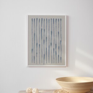 May include: A framed abstract art print featuring vertical blue brushstrokes on a neutral background. The artwork is displayed on a white wall above a table with decorative spheres and a wooden bowl.