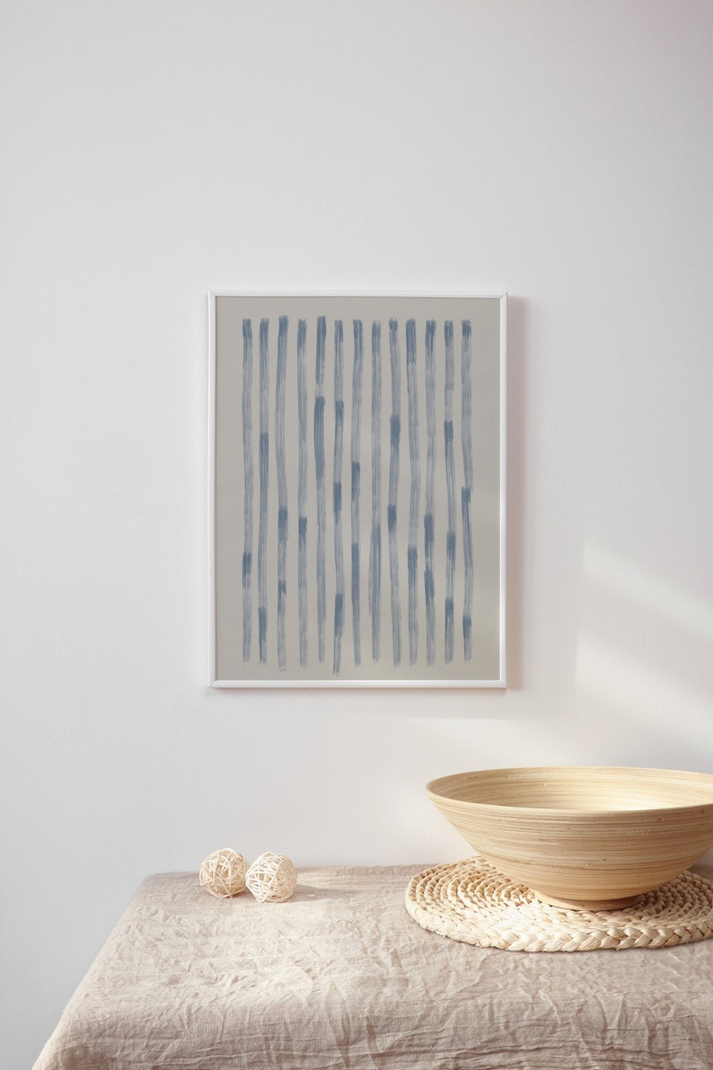 May include: A framed abstract art print featuring vertical blue brushstrokes on a neutral background. The artwork is displayed on a white wall above a table with decorative spheres and a wooden bowl.