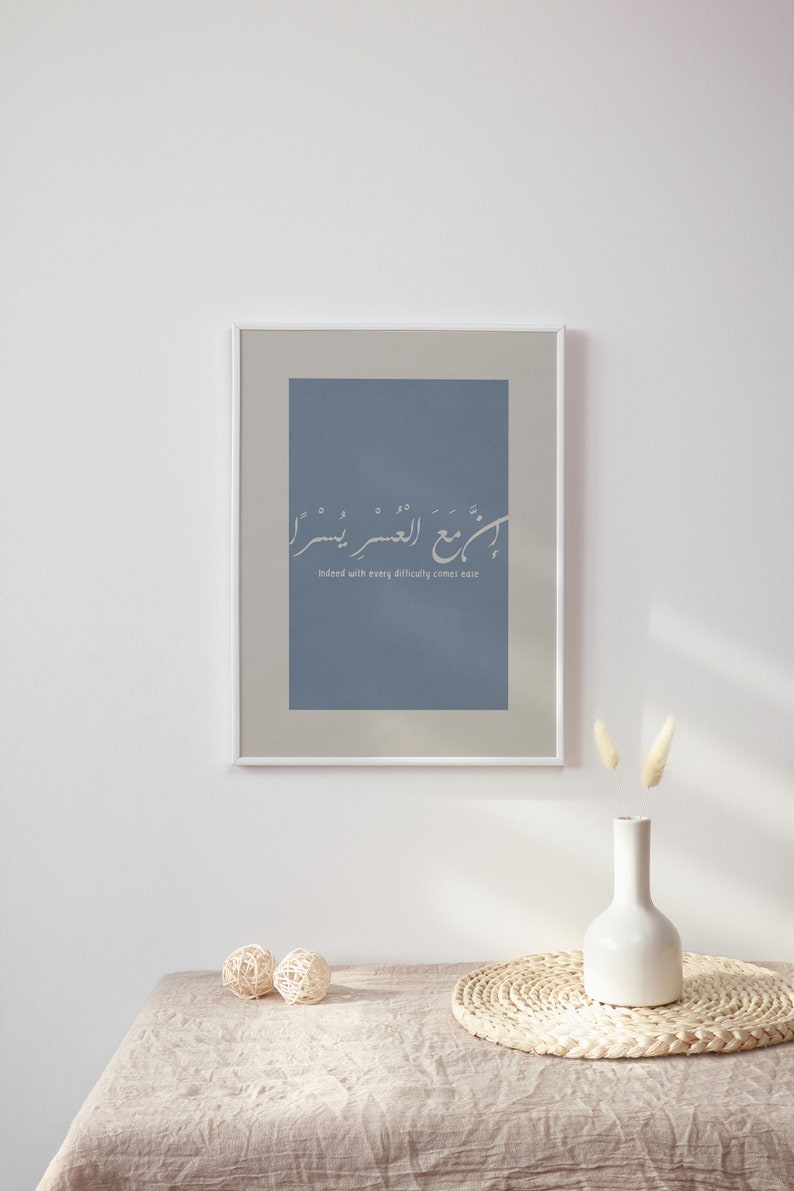 May include: Framed art print with Arabic calligraphy in white on a dusty blue background. The print is in a white frame and includes the English translation "Indeed with every difficulty comes ease". The artwork is displayed on a table with decorative elements.