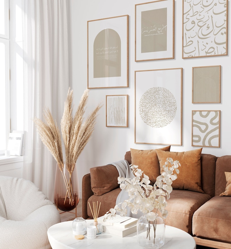 Op de afbeelding: Een woonkamerinterieur met een bruine leren bank, een witte fauteuil en een salontafel. De muren zijn versierd met een galerijmuur van ingelijste prints met abstracte ontwerpen en Arabische kalligrafie. Een vaas met gedroogd pampasgras en een vaas met witte bloemen staan op de salontafel.