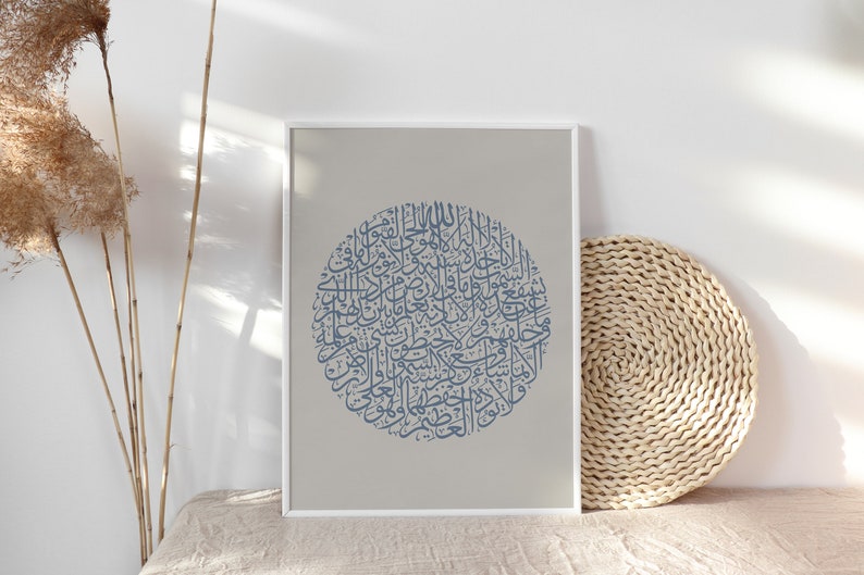 May include: A framed print featuring a circular design of Arabic calligraphy in a muted blue hue against a light gray background. The artwork is displayed on a neutral-toned surface, with dried plants and a woven mat in the background.