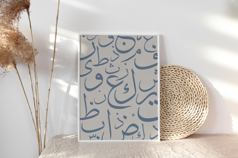 May include: A framed print featuring blue Arabic calligraphy on a light gray background. The artwork is displayed on a beige surface, with dried plants and a woven mat in the background. The print has a white frame.