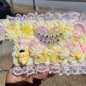 May include: A decorative phone case featuring a yellow and white frosting design with a heart that says "POM POM PURIN". The case includes a yellow cartoon dog, a pink moon, and a pink chocolate bar. The case is trimmed with pink lace.