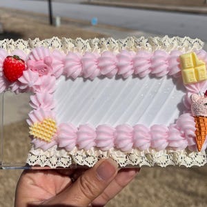 May include: A decorative license plate frame with a pink frosting design, adorned with a red strawberry, a bear-shaped figure, a waffle, and a chocolate bar. The frame has a white lace trim and a clear plastic base.