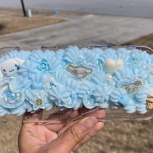 May include: A clear plastic case decorated with light blue frosting-like swirls, featuring cartoon character embellishments. The case includes a white heart, bows, and a small ice cream cone. The case is designed for a handheld gaming device.