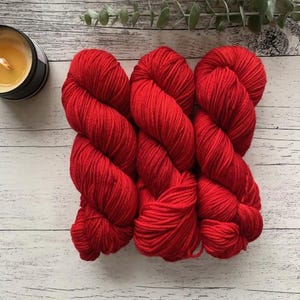 May include: Three skeins of bright red yarn are displayed on a light wood-effect surface. The yarn is tightly wound, showing the texture of the fibres. A small candle in a black container sits to the left, with greenery in the background.