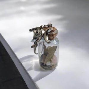 Könnte beinhalten: Eine kleine Glasflasche mit einem Korkverschluss und einem silbernen Anhänger. Der Anhänger zeigt einen Pentagramm und einen Halbmond. Die Flasche ist mit kleinen Stücken getrockneter Kräuter und Kristallen gefüllt.