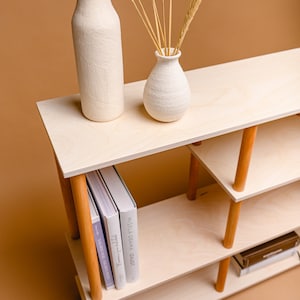 May include: A light wood shelving unit with three tiers, supported by wooden legs. Two white vases and dried wheat stalks are on the top shelf. Books are on the lower shelf. The unit is on a brown background.