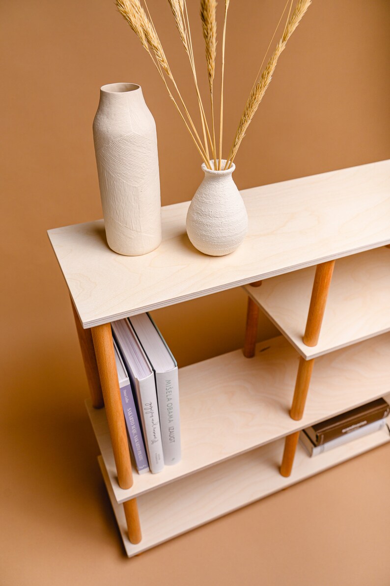 May include: A light wood shelving unit with three tiers, supported by wooden legs. Two white vases and dried wheat stalks are on the top shelf. Books are on the lower shelf. The unit is on a brown background.