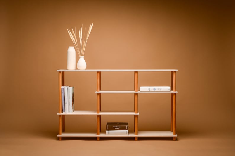 May include: A light wood bookshelf with three shelves and vertical wooden supports. The shelves hold books and decorative items, including two white vases and dried wheat. The bookshelf is approximately 122 cm wide.