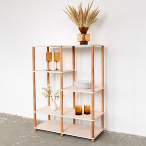 Puede incluir: Estantería de madera con cuatro estantes y soportes de madera. Los estantes exhiben objetos decorativos, incluyendo cristalería de color ámbar, un jarrón con flores blancas, un jarrón marrón con plantas secas y una pila de platos blancos.