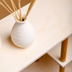 May include: A light-coloured wooden shelf unit with a white textured vase holding dried grasses. The shelf has two levels supported by wooden legs. The vase is round and has a textured surface.