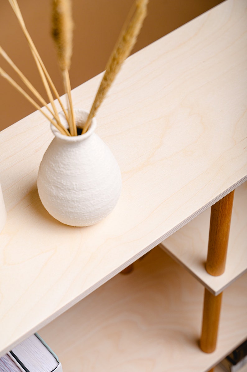 May include: A light-coloured wooden shelf unit with a white textured vase holding dried grasses. The shelf has two levels supported by wooden legs. The vase is round and has a textured surface.