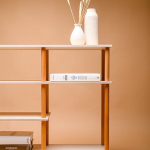 May include: A contemporary bookshelf featuring light wood shelves and wooden supports. The shelves display books and decorative vases with dried plants. The background is a warm beige colour.