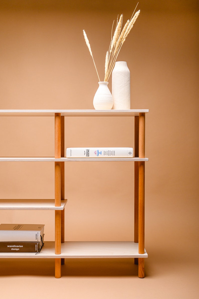 May include: A contemporary bookshelf featuring light wood shelves and wooden supports. The shelves display books and decorative vases with dried plants. The background is a warm beige colour.