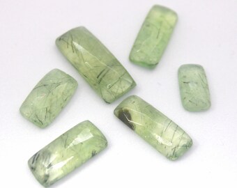 May include: Six green and black rectangular gemstone cabochons with a translucent appearance. The stones have a light green color with black inclusions that resemble thin lines or threads.