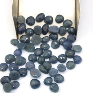 Könnte beinhalten: Ein loser Haufen blauer Saphir-Edelsteine, einige mit facettiertem Finish, die aus einer silbernen Schaufel auf eine weiße Oberfläche fallen.