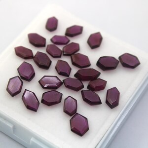 Könnte beinhalten: Eine Sammlung von 20 violetten Edelstein-Cabochons in einer weißen Schachtel. Die Cabochons sind facettiert und haben eine sechseckige Form.
