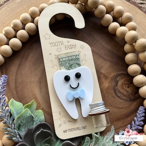 May include: Wooden tooth fairy door hanger with a white tooth cutout, blue bow, and a small glass jar. The door hanger has the text "Tooth Fairy" and "My Tooth Fell Out".